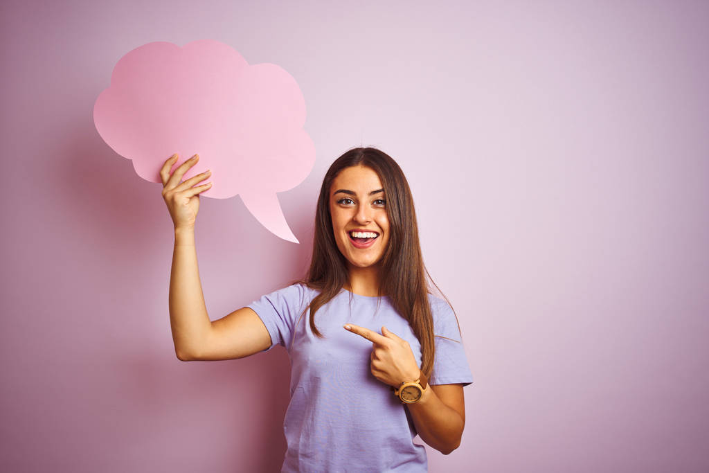 Young beautiful woman holding cloud speech bubble over isolated pink background very happy pointing with hand and finger